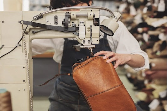 Person sewing a leather bag using a sewing machine in a workshop.
